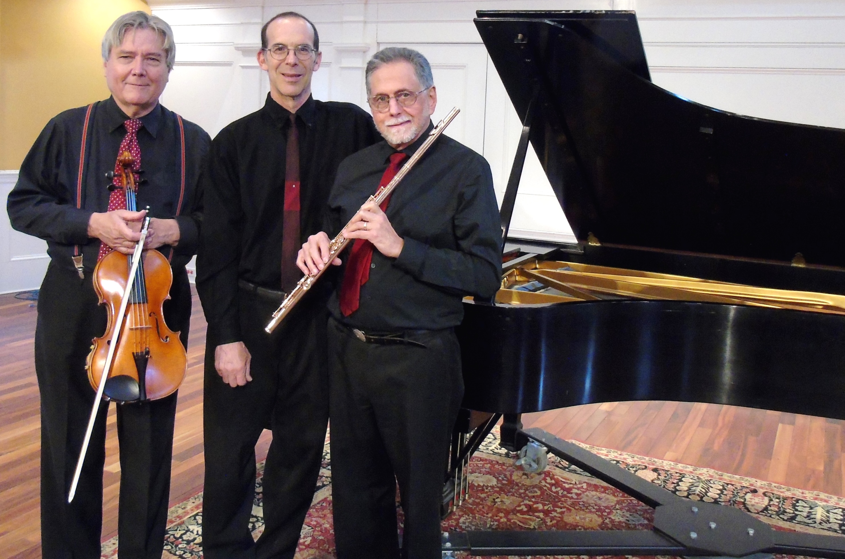 Three men wearing black dress shirts with red ties, the one on the left carries a violin, the one on the right carries a flute. They are all standing in front of a piano. 