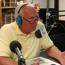 man in a yellow shirt that has blue headphones and a large microphone next to him. He is doing a radio show. 