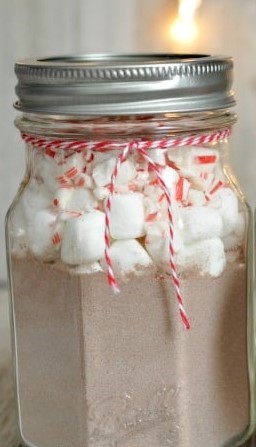 a mason jar filled with hot chocolate mix, marshmallows, and peppermint candies against a white background with a small yellow light. 