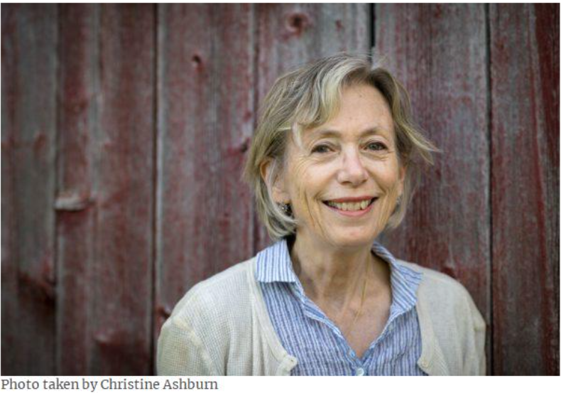 Photo of a person standing next to a faded red wood background. They are wearing a blue striped shirt and a cream colored sweater. They have blonde hair with a page boy haircut and earrings. They are smiling. The photo was taken by Christine Ashburn