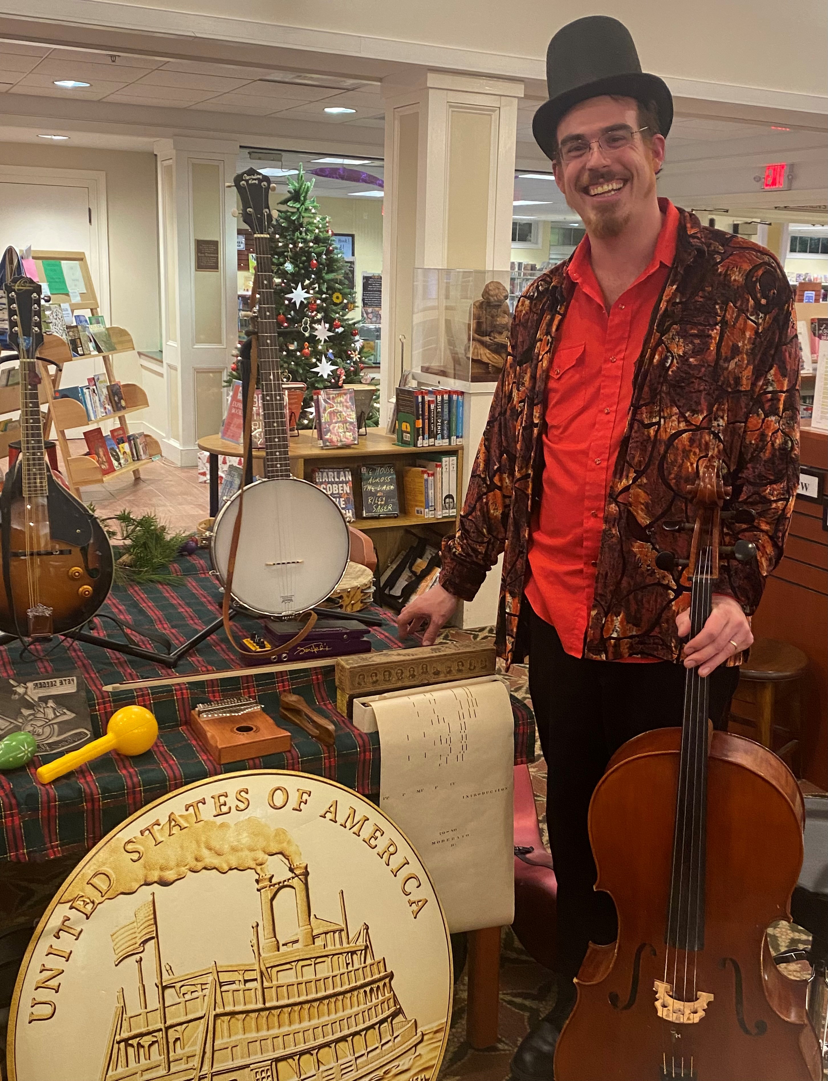 A gentleman in a top hat, velvet jacket and a red polo shirt stands next to several instruments on a table inside a library