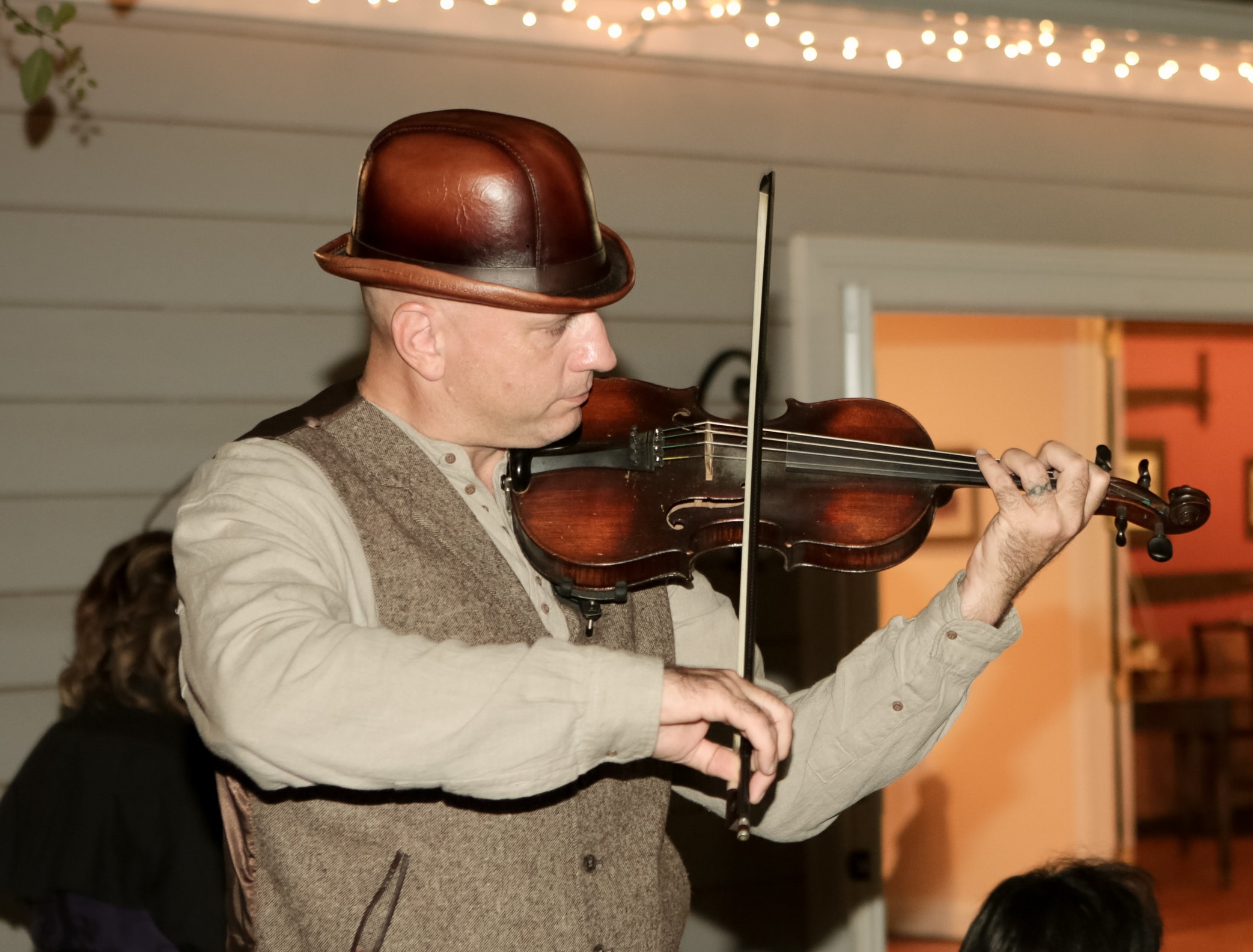 Person in a brown bowler hat playing an violin in a dark beige tweed jacket and a light beige undershirt. The background is of a private residence, side of the house. Picture appears to be candid.