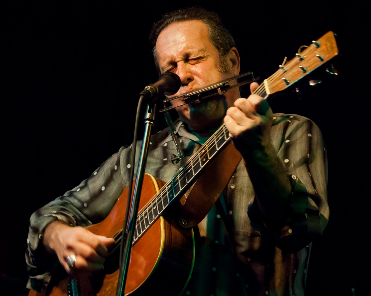 a man playing a guitar an a harmonica next to a microphone in a gray/black shirt with white dots against a black background. 