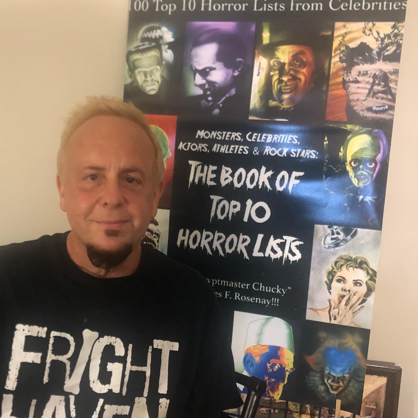 A white man in a black graphic t-shirt with blonde hair and dark pointed beard standing next to a stanchion display of his book