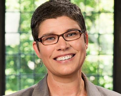 Jocelyn Kennedy, a woman with dark rimmed glasses, short hair, and a smile. She is against a blurred greenery background. 