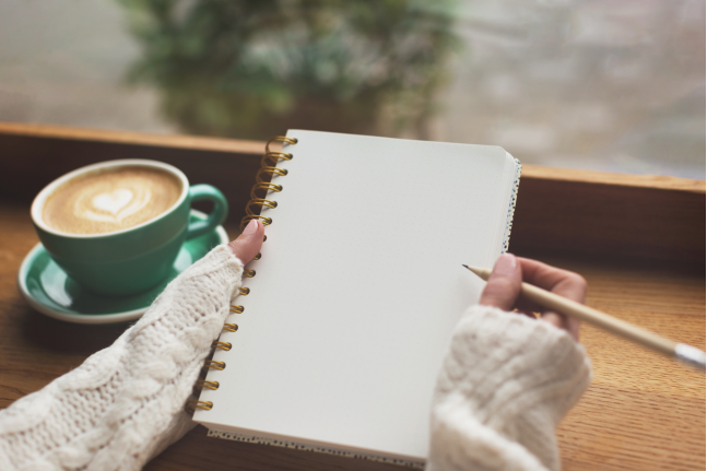 a person holding a spiral bound notebook at a wooden desk with a pencil and a latte with heart art on top of it in a green up. The person is a white knitted top and the rest of the of image is blurred. 