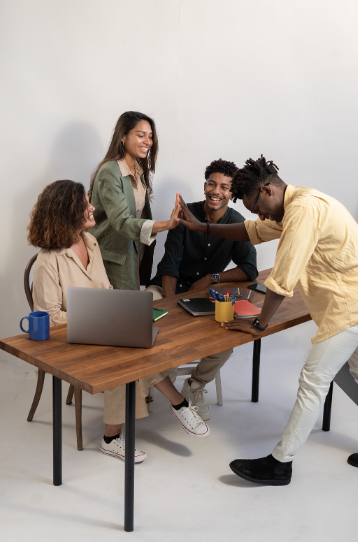 four people working together at a wooden desk with a laptop and some writing utensils. One of them is highfiving the other and they all seem to be happy and smiling.  