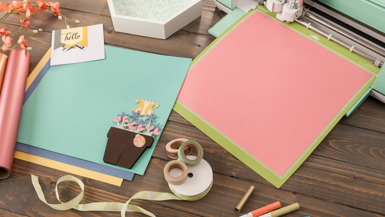 a collection of paper material that is against a dark wooden background that has a cricut machine kitty cornered in the upper corner. There is also ribbon and some red flowers in the space.