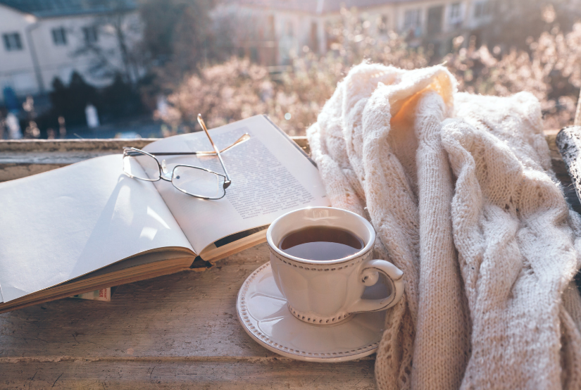 a open book with glasses on top of it next to a cup of tea and a off-white blanket 