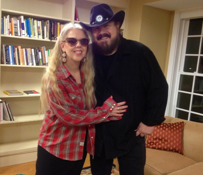 two people, slightly embracing, one presenting woman, one presenting man, one has long blonde slight curly hair and one is dressed in mostly black with a black cowboy hat 