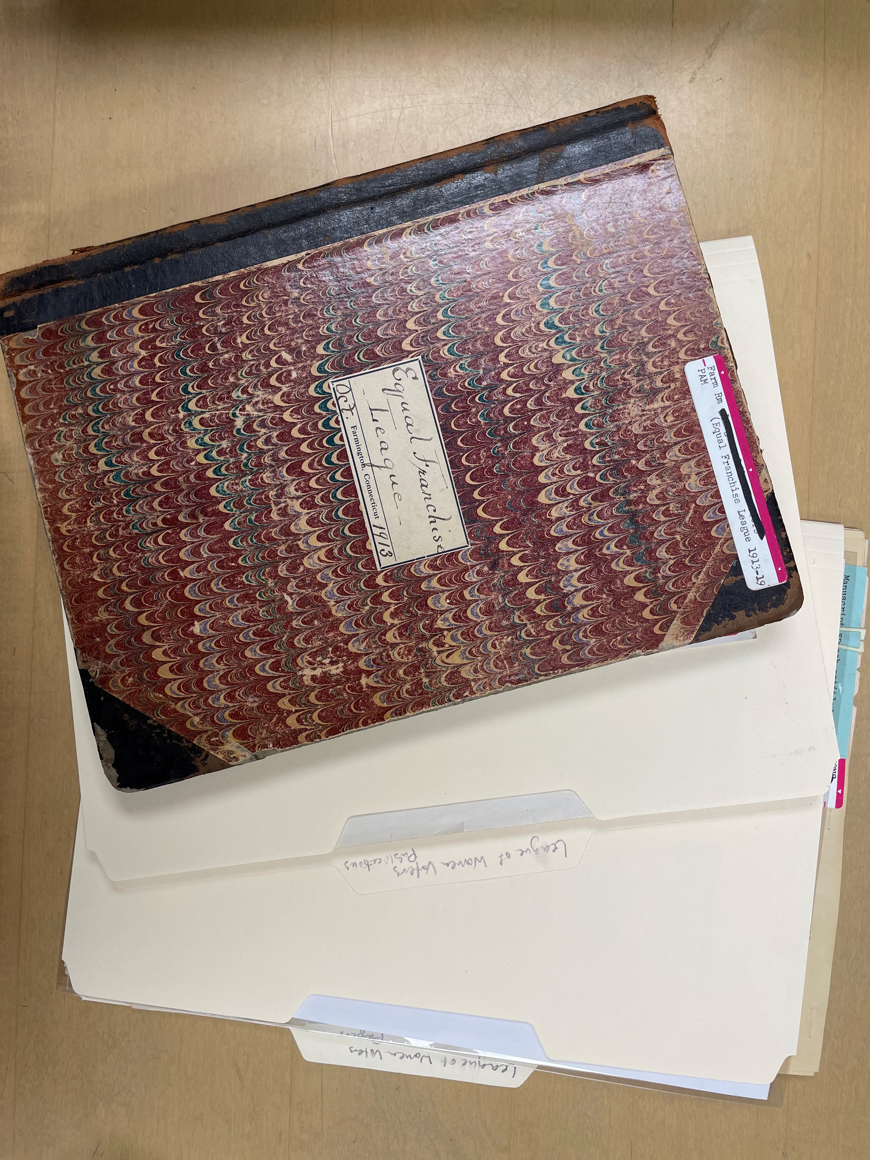 A record book with a marbled cover and two file folders fanned out on a tabletop. On the front cover of the record book is written by hand, “Equal Franchise League, October 1913.