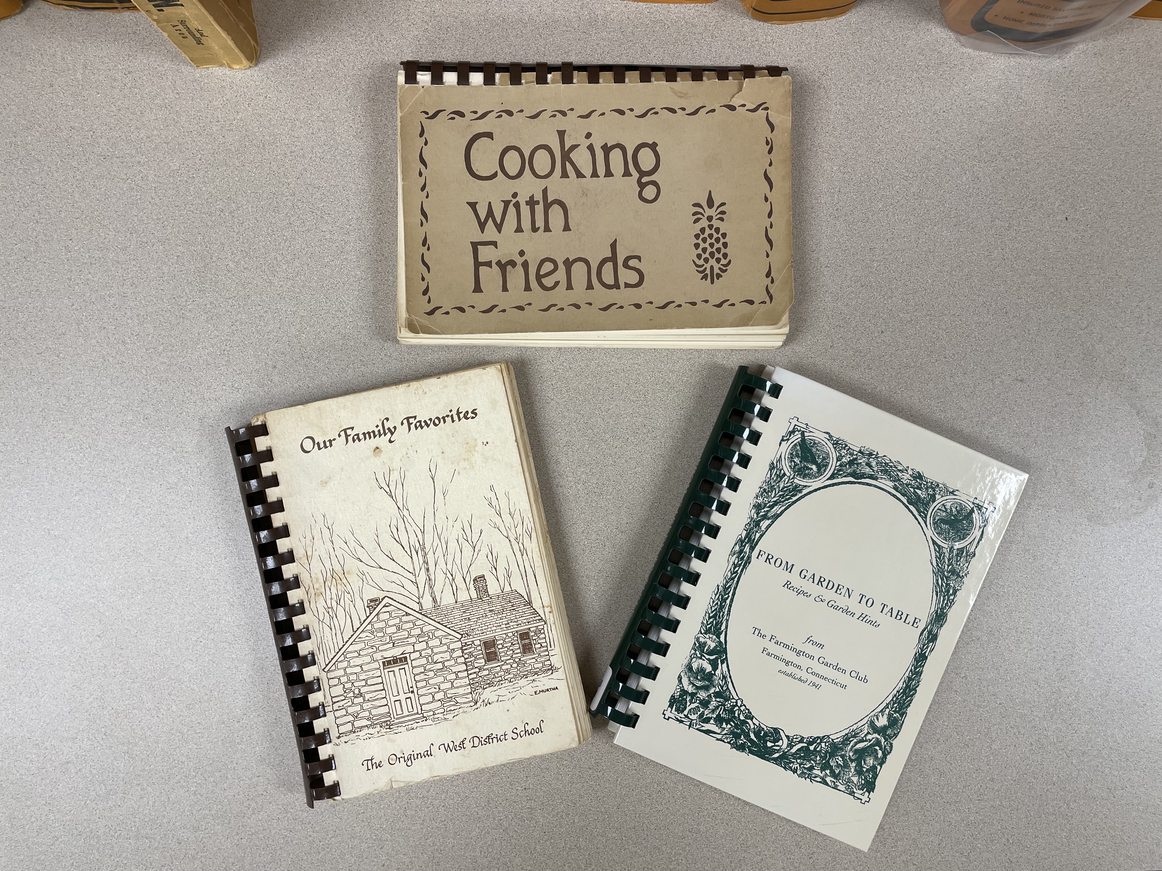 Three spiral-bound cookbooks on a counter, arranged with the covers clearly visible. Clockwise from top: Cooking with Friends, a light brown card stock cover with stencil-style graphics in dark brown ink; From Garden to Table, a light green, glossy hard cover with dark green lettering and graphics; Our Family Favorites, a cream-colored card stock cover with brown lettering and an illustration of “The Original West End School.”