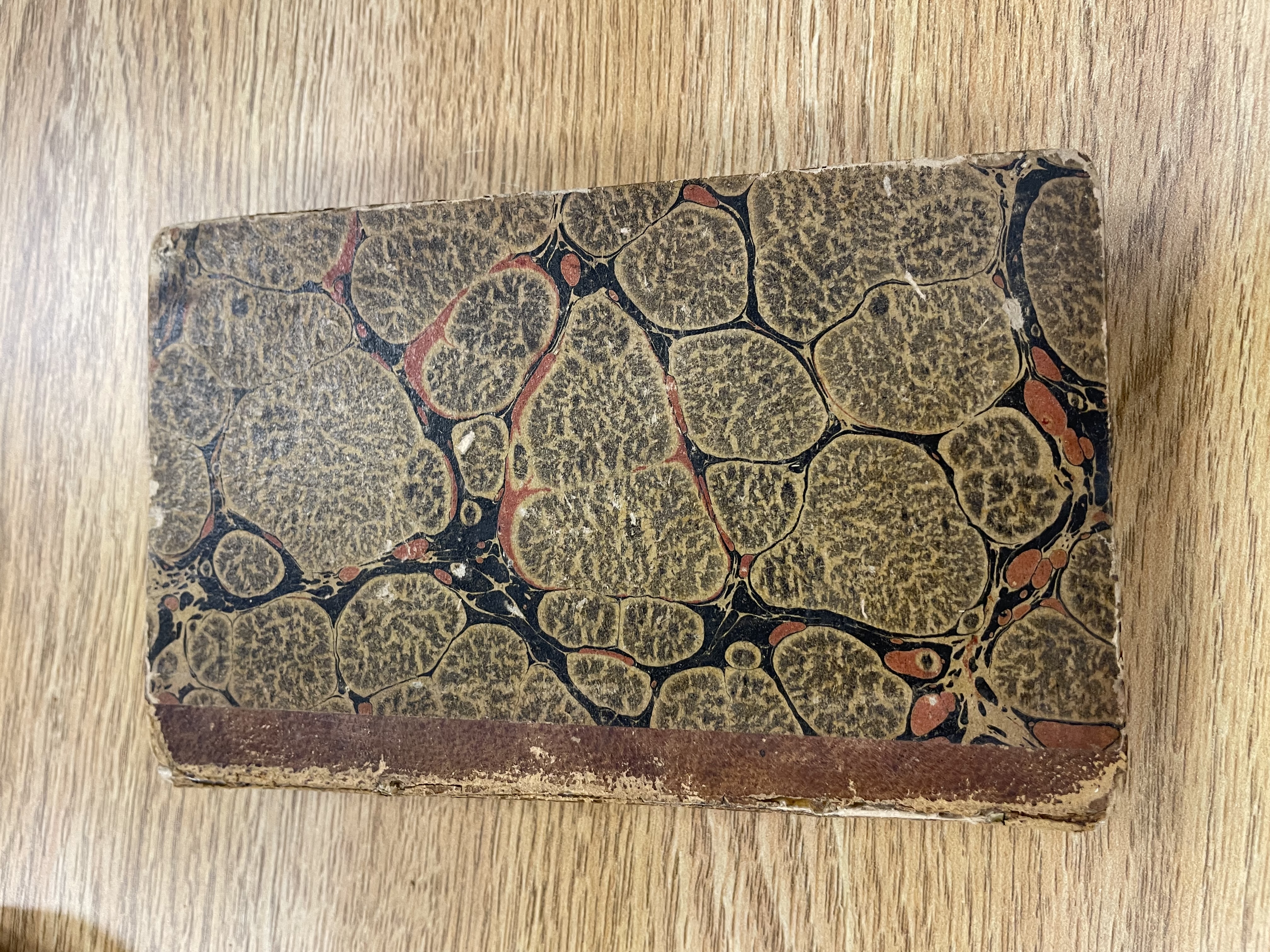 A small, old book with leather spine and a front cover marbled with black, red, and light brown. The marbling pattern is reminiscent of cells under a microscope.
