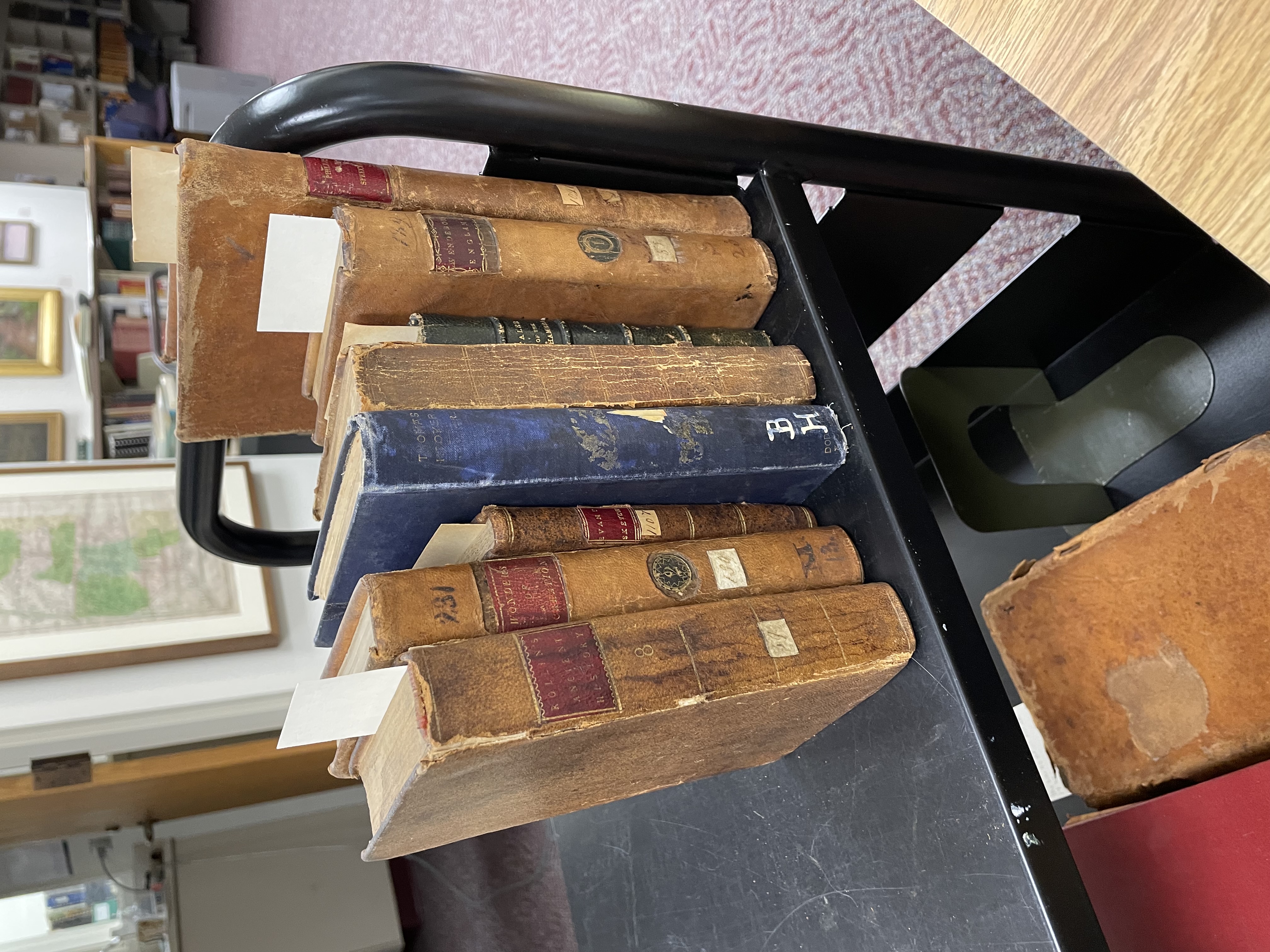 Eight old books of various sizes sitting on a metal cart. Some have numbers on the spines; most have leather covers.