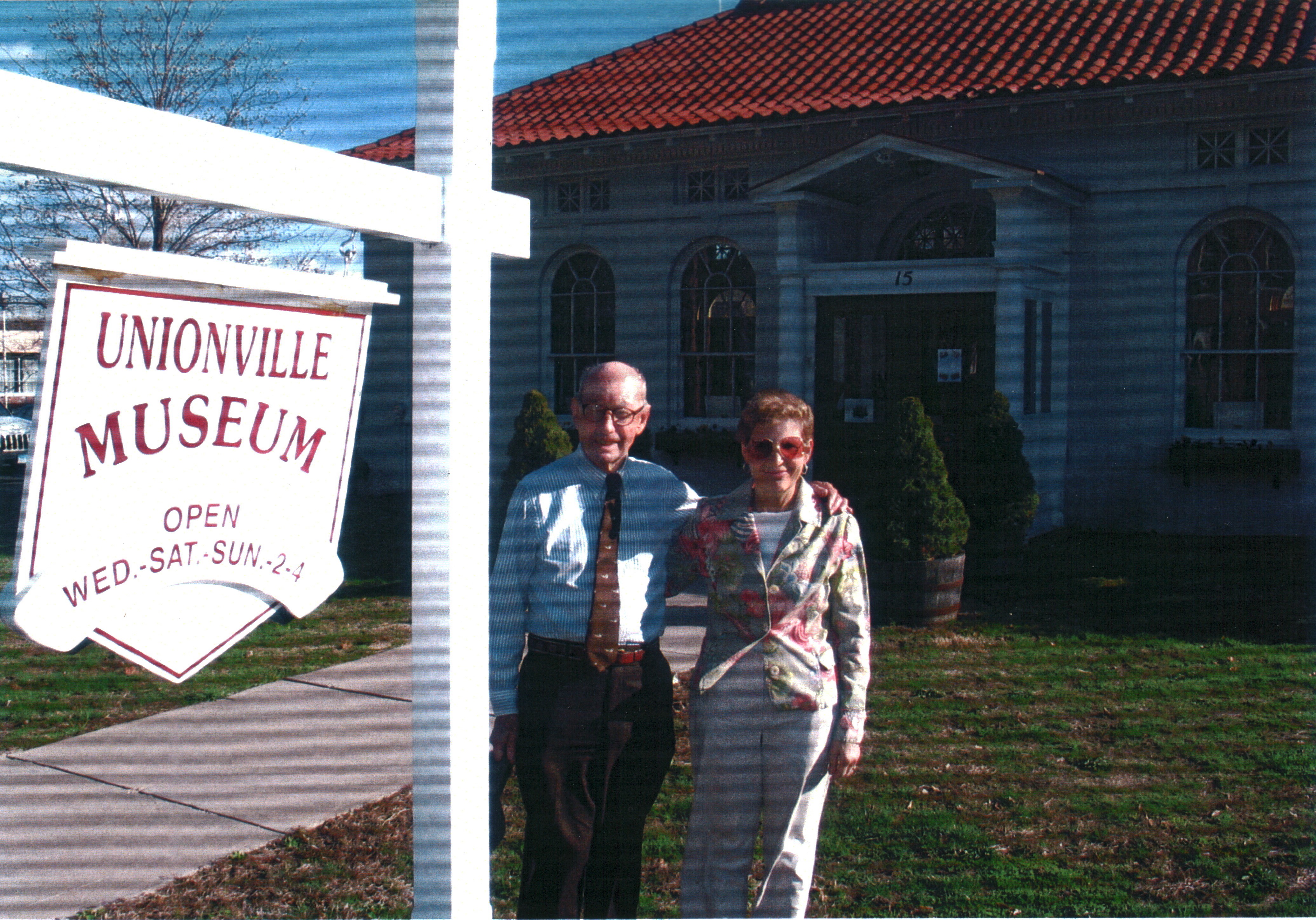 Two adults stand with their arms around each other's shoulders, smiling. They stand in front of a small, early 20th century library building and next to a white sign with maroon lettering that says “Unionville Museum, Open Wed.-Sat.-Sun.-2-4"