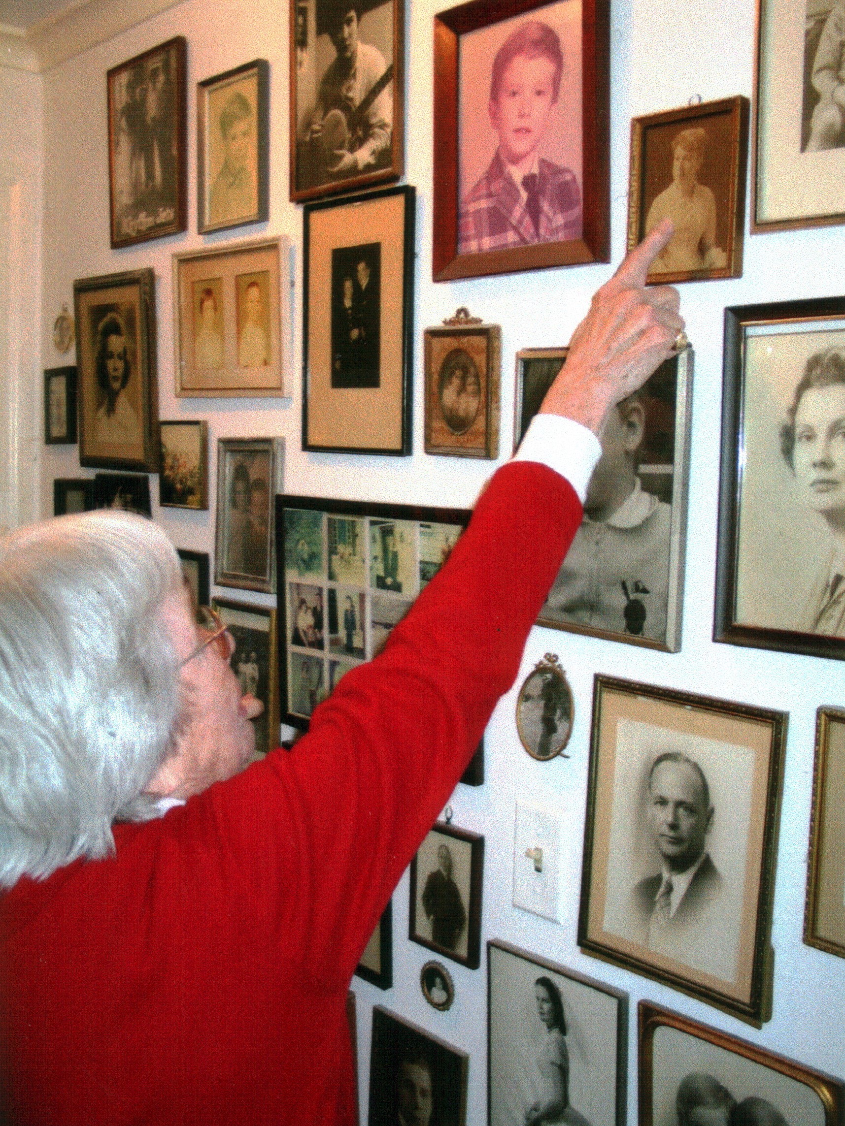 A profile of a woman with silver hair and glasses, wearing a red sweater, facing a wall full of framed photographs of family members. She points to a particular photo high on the wall.