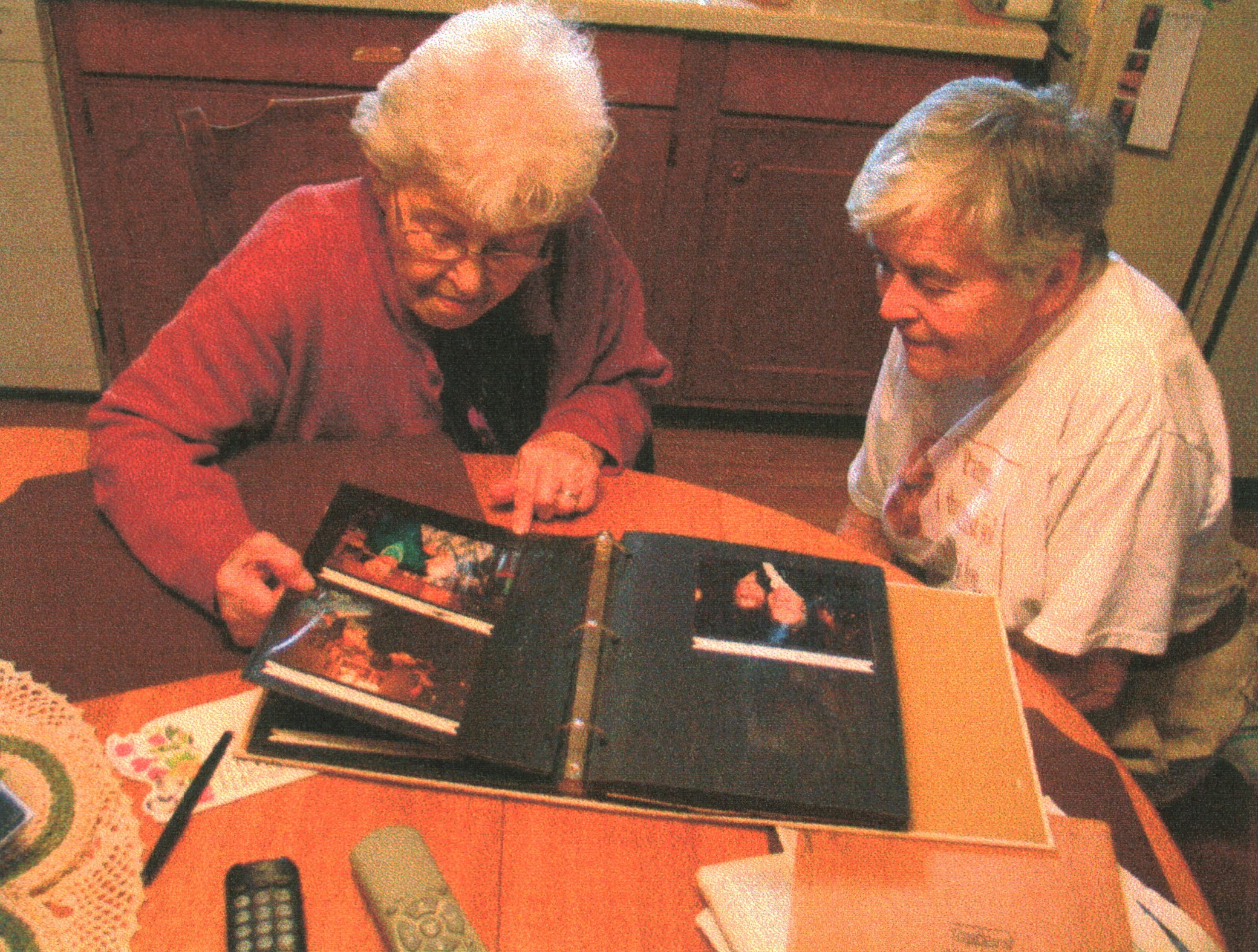 Two women are sitting at a kitchen table. A photo album is open on the table. One woman is pointing out a particular photo while the other woman looks on.