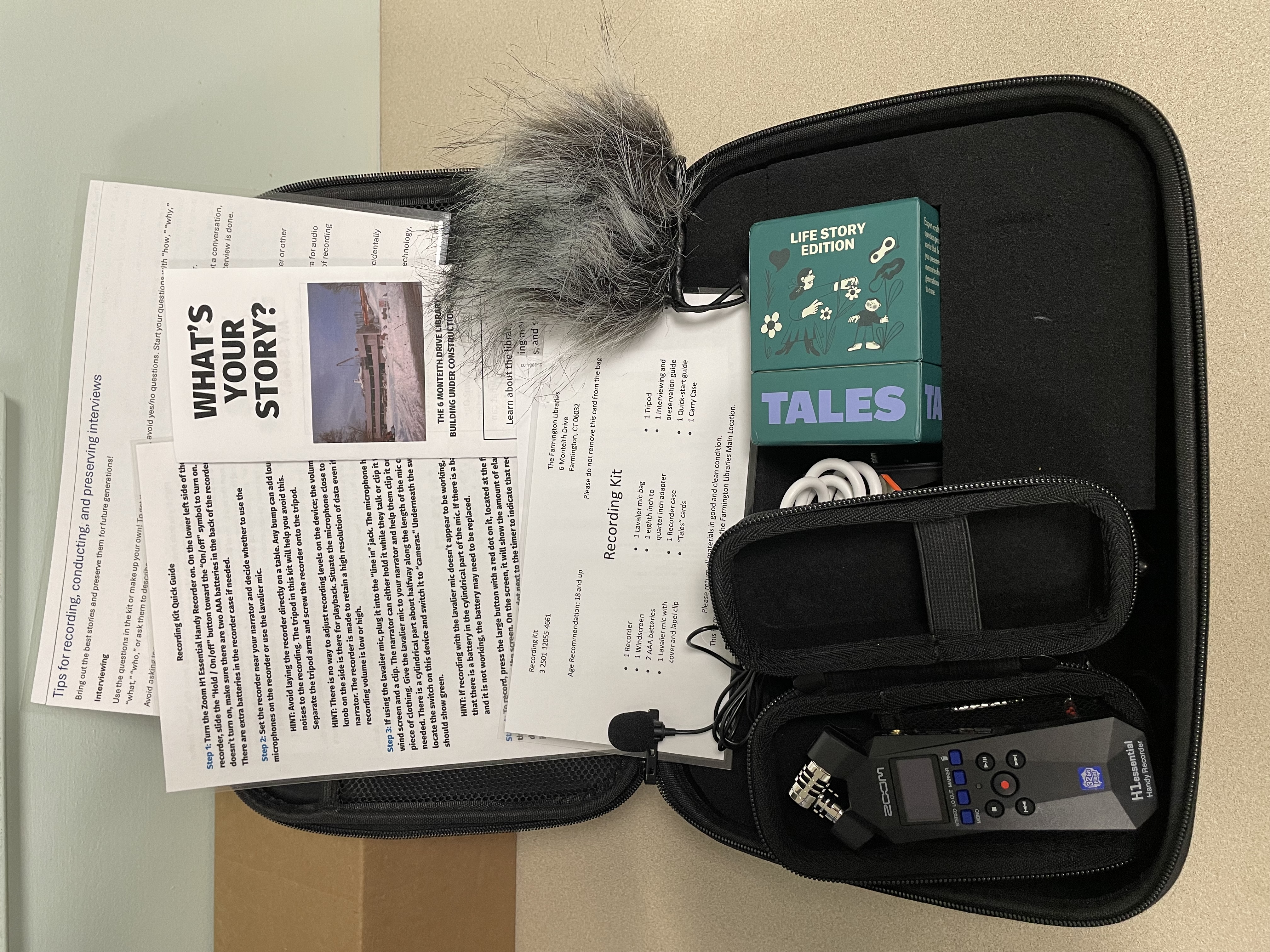 An open case on a table with laminated guides, a handheld recorder, a furry windscreen, a green box of cards, and a lavalier microphone.