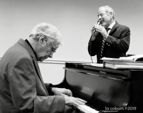 a black and white image of two men, one playing piano and the other singing into a microphone. Both are wearing suits.