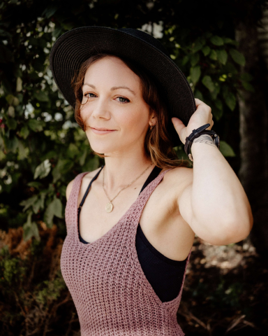 a woman in a mauve shirt against a greenery background holding onto her hat. She is looking into the camera from a 45 degree angle. 