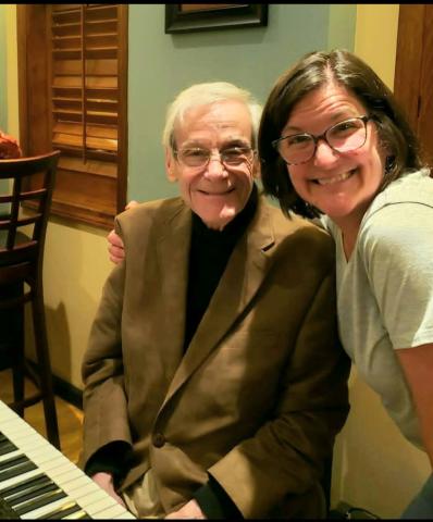 two people smiling into the camera. One is seated at a piano while the other is standing with their arm around the seated person.