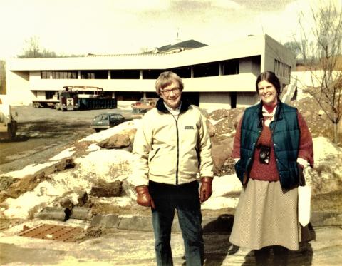 [alt text] A man and a woman stand smiling in front of a library building and parking lot under construction in 1983. Immediately behind them are piles of dirt and gravel. A large dumpster and work truck sit in the parking lot near the front entrance. Several cars are parked in front of the building. It is a partly cloudy day, possibly in early spring.