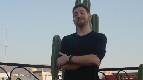 A man stands on a rooftop terrace with arms crossed, wearing a black shirt and bracelets. Tall cacti are behind him, with buildings and a clear sky in the background.