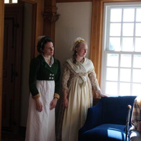 Two women in historical 18th–19th century clothing stand by a window. One wears a green jacket over a white dress; the other wears a cream gown with a shawl. Sunlight streams in as they look outside, near a blue armchair.