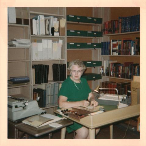 A woman with gray hair and wearing a green dress sits at a desk, looking at the camera. Around her are shelves containing archival boxes and books. On the desk is a typewriter, a small lamp, a set of rubber stamps, and several archival items being processed. Inscription on the back reads, “Alice B. Cushman Cataloging the Farmington Room, September 1967.”