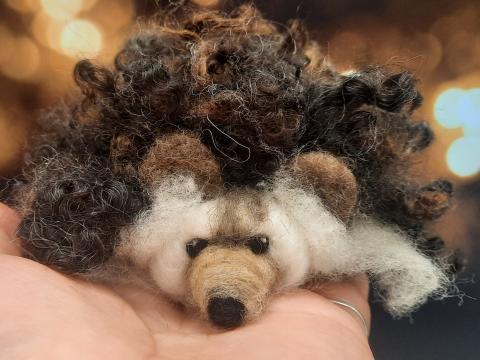 A small, handcrafted hedgehog made of needle-felted wool rests in the palm of a person's hand. The hedgehog has a cream-colored face and belly, a brown nose, black eyes, and short rounded ears. Its back is covered in thick, curly dark brown and black wool, mimicking spiky fur. The background is softly blurred with warm golden lights.