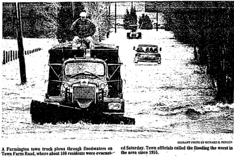 Black and white photo of a large town truck driving through deep floodwaters on a road. A man sits on top of the truck cab as it pushes through the water. Behind it, other vehicles are partly submerged as they follow through the flooded street.