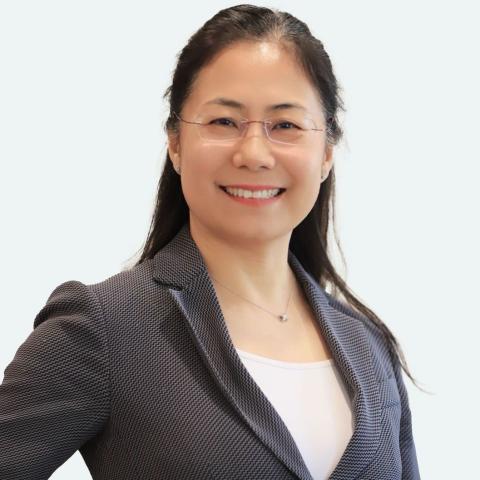 A female smiling and looking directly at the camera with long black hair that has been tied back. She is against a white background and wearing a grey patterned jacket.