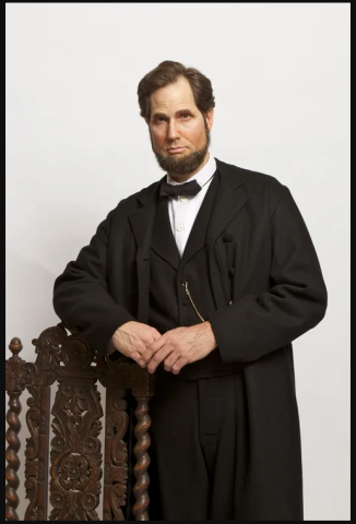 a Lincoln impersonator that is leaning against a dark wooden chair against a stone colored background 