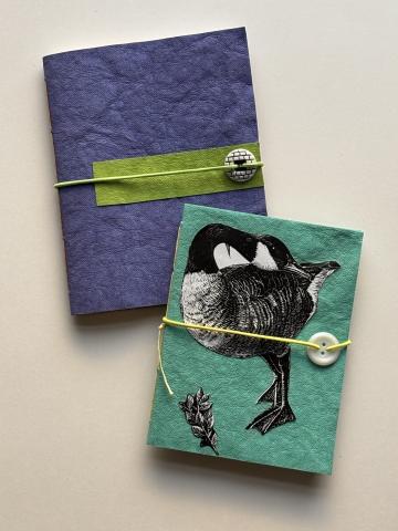 a photo of two handmade journals with button clasps. they are both face up to show the front cover. The top one is dark blue with a green accent and the bottom one is teal with a goose on the front. 
