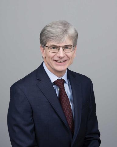 an older man in a blue suit and burgundy tie against a gray background