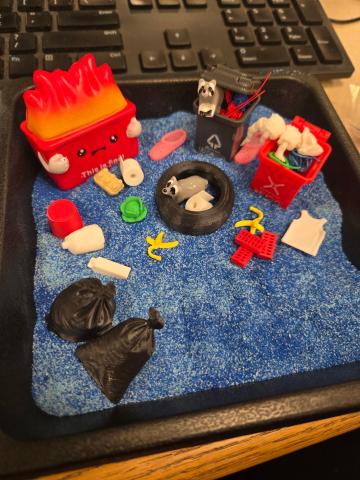 An assembled Trash garden, showcasing a dumpster on fire alongside raccoons, opposums, and various trash items.