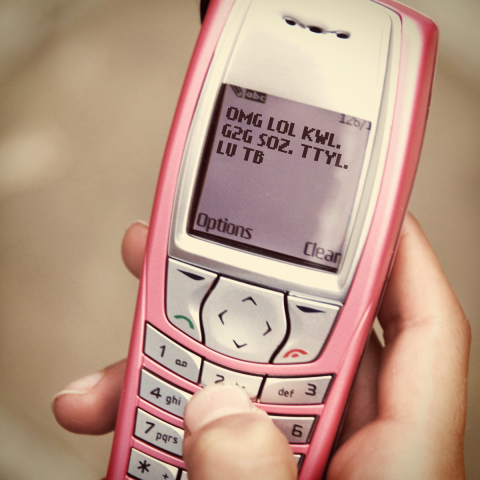 A pink "early" cell phone, held by a hand texting out the phrases: "OMG LOL KWL! G2G SOZ. TTYL. LU TB."