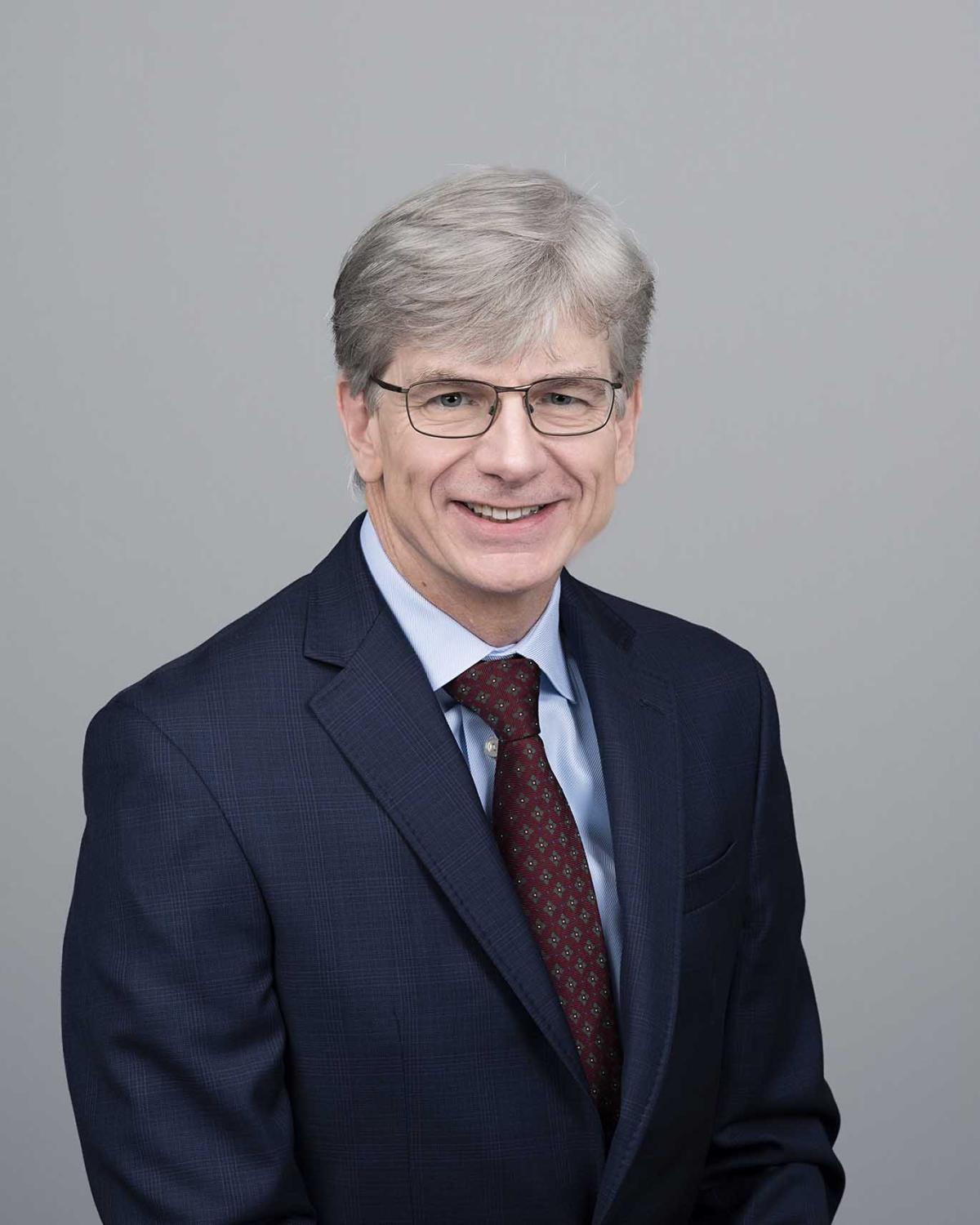 an older man in a blue suit and burgundy tie against a gray background